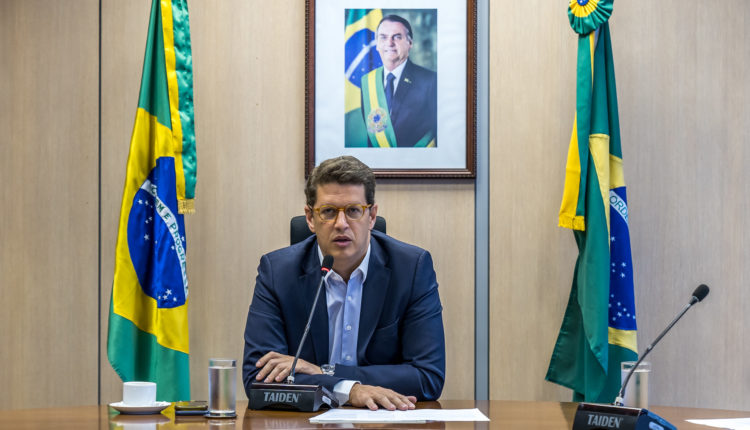 Ricardo Salles, Ministro do Meio Ambiente de terno azul sentado em uma mesa com bandeiras do Brasil ao fundo.